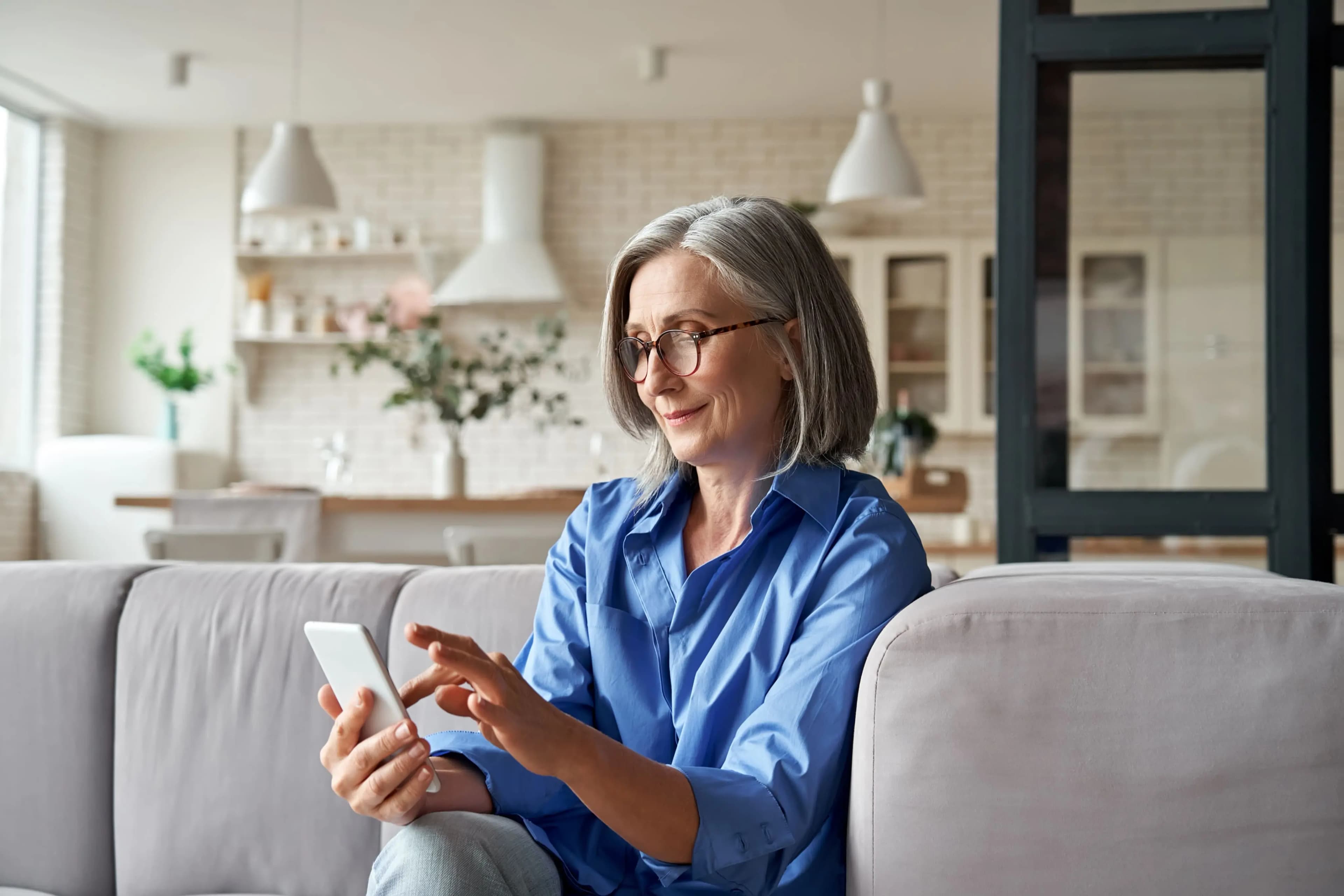 A woman with gray hair and glasses is sitting on a couch using a smartphone in a well-lit living room.