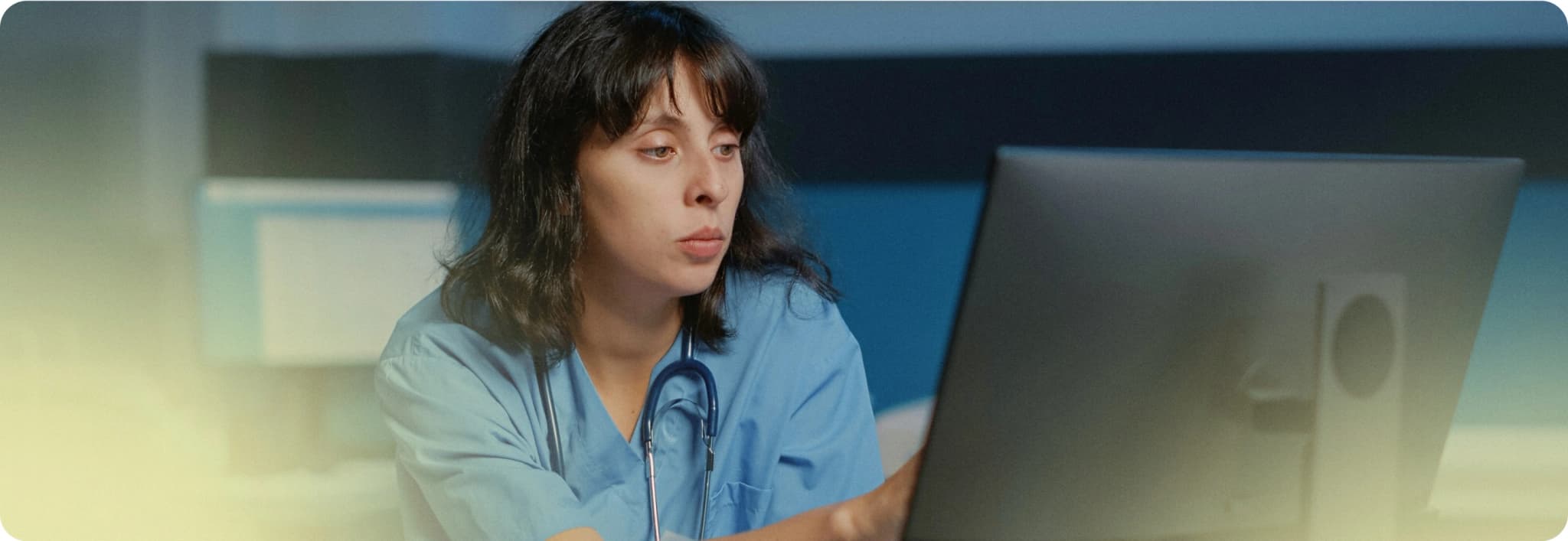 Person in scrubs sitting at a desk with a computer monitor, in a medical office setting.